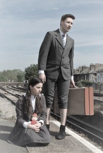 A girl crouches on a railway platform while a boy stands behind her