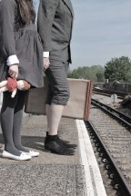 A boy and a girl stand on a railway platform, clutching a doll and suitcase. Their heads are not visible.